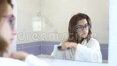戴眼镜的年轻女子在镜子前梳头。 一个穿着白色浴袍的女孩在浴室镜子前