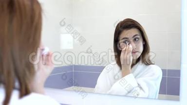 一个年轻的女人用棉垫从脸上卸妆。 一个穿着白色浴袍的女孩在浴室镜子前