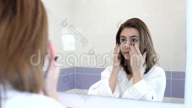 一个年轻的女人在镜子前把面霜涂在脸上。 水疗程序。 <strong>浴</strong>室里穿着<strong>白色浴袍</strong>的女孩
