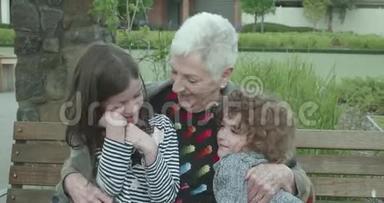 当两个小孩坐在公园的长凳上拥抱时，一位年长的妇女拥抱并挤压着他们，而这个小男孩正拥抱着他的奶奶
