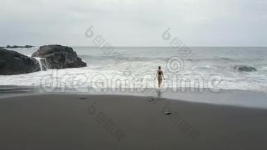 在西班牙特内里费的一个黑色沙滩上看到一个女人在水中奔跑。 海浪<strong>冲破</strong>，形成白色