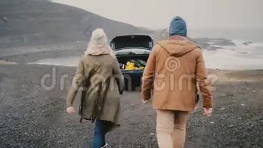 年轻快乐的一对坐车旅行。 迷人的男人和女人走到冰岛黑色海滩附近的车辆。