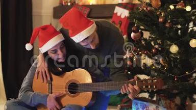 <strong>圣诞</strong>前<strong>夜圣诞</strong>老人戴`帽子的幸福夫妇的肖像。 男人教女人弹吉他。 <strong>圣诞</strong>树上的<strong>圣诞</strong>树