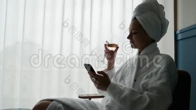 优雅的女人穿着<strong>白色浴袍</strong>，头上有一条毛巾，享受着一杯葡萄酒，在酒店里使用手机