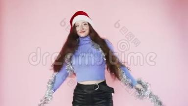 美丽的红发女孩戴着圣诞老人的帽子旋转。 新年的心情。 圣诞节。 慢动作。 在粉色背景上。