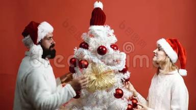 男人和女人用红色背景上的红色球装饰白色圣诞树。 爱的情侣准备新年晚会