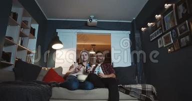 快乐的年轻，放松的白种人，浪漫的夫妇笑着在家看有趣的电影，用投影仪慢动作吃零食。