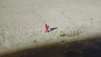圣诞老人站在湖边的沙滩上，手里拿着一个礼品袋。 圣诞老人在镜头前打招呼和<strong>打手</strong>势