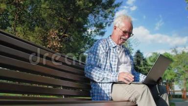 快乐老人在Skype上交谈，坐在花园的长凳上。 一个养老金领取者把笔记本电脑放在膝上。 他是