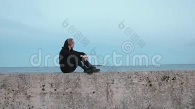 一个穿着黑色衣服的年轻人的肖像，他坐在靠海的码头上放松