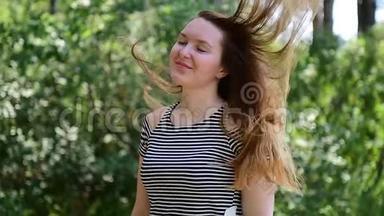 年轻的棕色头发的女人，穿着条纹t恤，站在阳光下，迎着绿色的<strong>树叶</strong>，风<strong>吹</strong>起她的头发。 漂亮姑娘