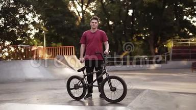一个带着BMX自行车的年轻人站在他的自行车旁边，旁边有斜坡，背景上有溜冰公园。 BMX骑手画像