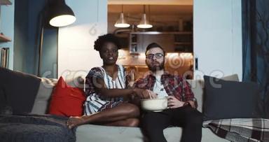 快乐的年轻白种人男人和非洲女人坐在家里一起看戏剧电影，吃爆米花慢动作。