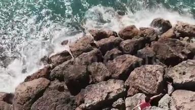 穿红色衣服的女人站在岩石悬崖上，双手分开，蓝色的水<strong>击打</strong>在岩石码头海滩上。 鸟瞰图