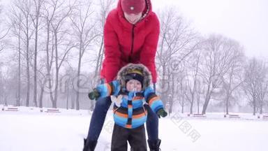穿着红色羽绒服的年轻女子在冬天的城市公园背景下，在雪地里缓慢地把她的小儿子卷在冰上
