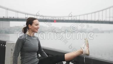 <strong>喜笑颜开</strong>的女人穿着活动服装在城市的堤岸上伸展，背景是混凝土桥。 年轻女运动员担架