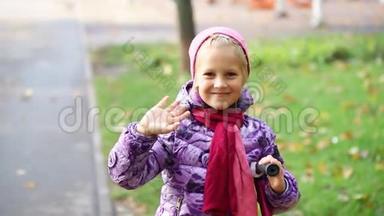 可爱可爱的白种人金发快乐的学校女孩微笑和挥手在城市公园温暖的秋天。 快乐的孩子