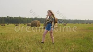 乡下女孩穿着格子衬衫和牛仔裤，在农田里摆姿势。 干草堆背景的牛仔女孩