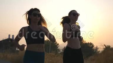 两个运动的女人沿着乡村道路慢跑，背景是日落。 晚上在户外工作的年轻女孩