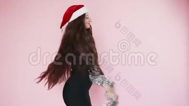 美丽的红发女孩穿着圣诞老人的帽子旋转和黑色的连衣裙。 新年的心情。 圣诞节。 慢动作。 在粉色背景上。