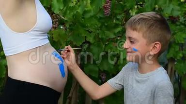 笑的男孩用刷子在他怀孕妈妈宝宝的肚子上画`脚印。 怀孕的概念。 特写
