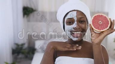 美丽的年轻的美国黑人妇女头上戴着白色毛巾敷面膜，手里拿着半个葡萄柚