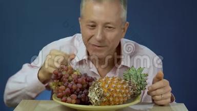一个穿着衬衫的中年男人慢慢地享受着吃葡萄的乐趣，他躺在一个大盘子里，放着菠萝，微笑着。