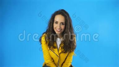 穿着黄色夹克的年轻女人在蓝底工作室<strong>咬</strong>着<strong>嘴唇</strong>和眨眼睛。