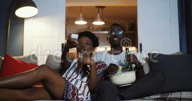 快乐年轻积极的黑人朋友，男人和女人看电视，在家里用遥控器拿<strong>零食</strong>和<strong>饮料</strong>慢动作。