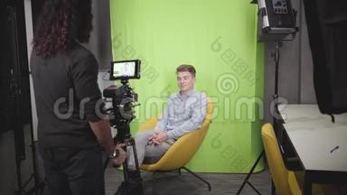 年轻的积极的白人男子坐在黄色的扶手椅上，在镜头前交谈。 摄像师录制视频.. 成功