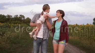 一家人带着一个小宝宝，带着向日葵在田野里走来走去，笑着。 农业。 女孩，母亲和父亲