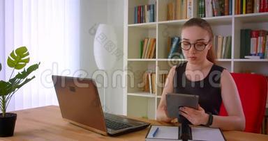 年轻漂亮的红头发女商<strong>人</strong>戴着眼镜，在办公室里用平板<strong>电脑坐</strong>在笔记本<strong>电脑前</strong>