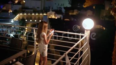 少女抱着手机在夜阳台.. 年轻女孩在户外露台上用智能手机点亮灯笼