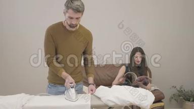 成年白种人<strong>男子</strong>熨烫<strong>深色</strong>头发的女人坐在后台。 丈夫在妻子休息时做家务