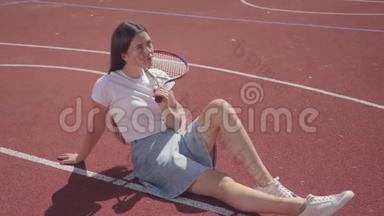 穿着运动鞋、裙子和T恤的漂亮黑发<strong>少女</strong>坐在户外篮球场上，手里拿着<strong>网球</strong>
