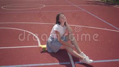 穿着运动鞋、裙子和T恤的漂亮黑发少女坐在户外篮球场的滑板上。