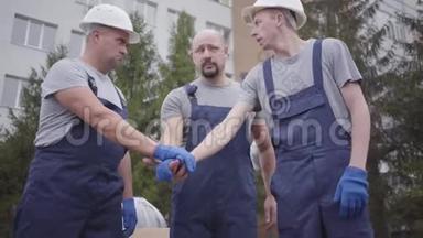 三名白人青年工人互相握手，摇头，开始工作。 两辆白色头盔的搬运车