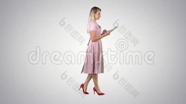 金色的粉红色连衣裙行走和使用平板在渐变背景。