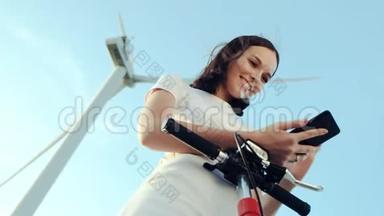 这个女孩在她的<strong>电动</strong>滑板车旁边使用一部智能手机，背景是风力发<strong>电机</strong>。
