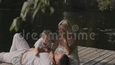 年轻幸福的家庭，爸爸妈妈和儿子在河边的码头上阳光明媚的一天。 男人躺在女人腿上