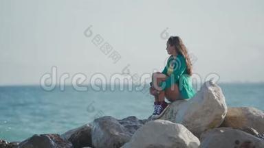 美丽的女人穿着运动鞋和夹克，坐在岩石海滩上，望着海浪拍打着岩石