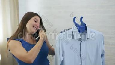 一个年轻的女人在电话里说话，用蒸汽<strong>抚摸</strong>男人`衬衫。 蒸衣服蒸的过程.. 蓝色