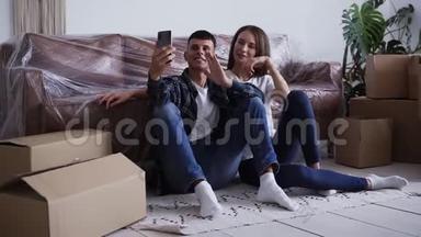 年轻快乐的白种人夫妇坐在他们的新房子的地<strong>板</strong>上，自拍或给他们的朋友<strong>打视频</strong>电话