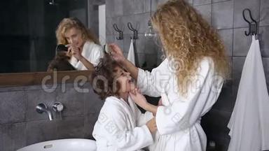可爱的女人穿着白色浴袍，梳着卷发，她的小儿子在家里的浴室里。 <strong>每天</strong>的卫生，快乐