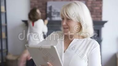 一幅成熟的女人坐在沙发上看着平板电脑上的照片，而她的女儿则在打扫房间。 妇女