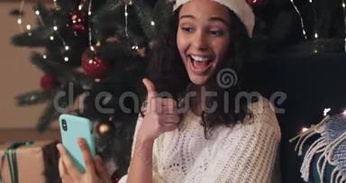 圣诞老人帽子里的年轻女孩坐在装饰好的圣诞树下，用她的智能手机进行视频聊天