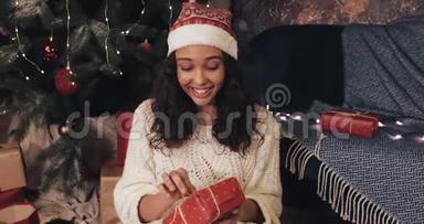 美丽的微笑<strong>穆拉托</strong>女孩坐在圣诞树下舒适的家背景，同时解开红色礼物盒