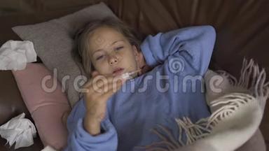 生病的年轻白种人女孩在家里躺在毯子下面用温度计测量体温。 孩子有