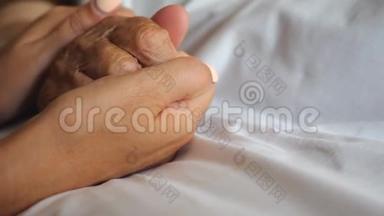 女人牵着她的老母亲躺在床上，表现出关怀或爱。 女儿安慰老妈妈布满皱纹的手臂