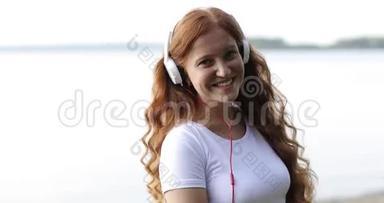 把脸贴紧。 带着白色耳机的红发雀斑女孩正在湖畔听歌跳舞。